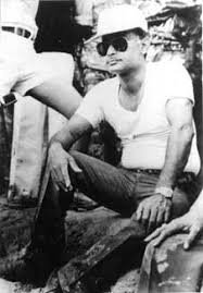Ziaur Rahman seated outdoors in a white shirt, dark trousers, sun hat, and sunglasses at a field site.
